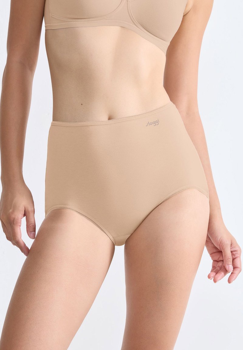 Kvinna som bär högmidjade beige underkläder och matchande behå, stående mot en enkel ljus bakgrund, med armarna avslappnat vid sidorna.