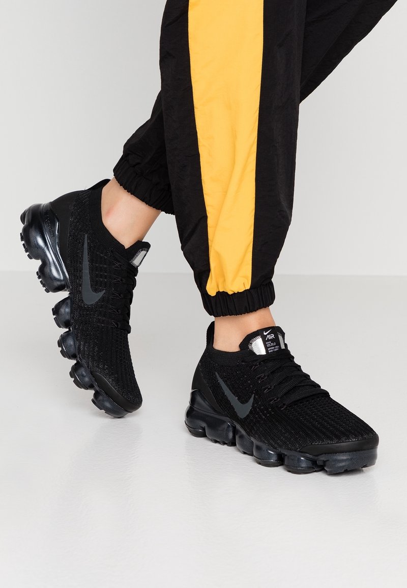 Svarta stickade sneakers med en strukturerad ovansida, svart gummisula och grå Nike-logga. I kombination med svart och gulrandiga joggers.