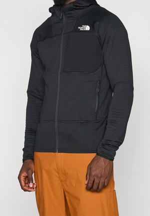 Man wearing black zip-up hoodie with chest pocket and white North Face logo, paired with orange pants, standing against plain background.