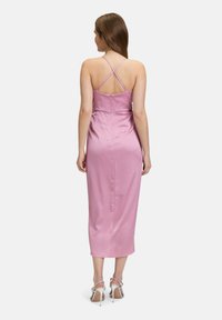 Robe en satin rose avec une silhouette cintrée, longueur midi et bretelles croisées dans le dos. Associée à des chaussures à talons hauts argentées.