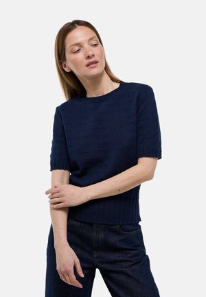 Femme portant un pull en maille bleu marine à manches courtes et un jean foncé, les bras légèrement croisés, regardant légèrement sur le côté.