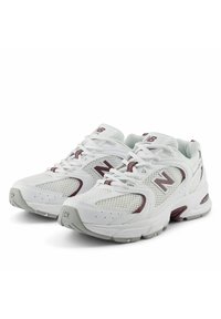 Scarpe da ginnastica bianche con pannelli in rete, dettagli rossi e linguetta imbottita. Presentano suole in gomma robuste e il logo "N" sul lato.