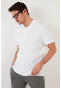 Camisa polo blanca de canalé con cuello clásico, que presenta mangas cortas y una tapeta de tres botones, acompañada de pantalones gris con rayas.