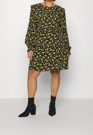 Kobieta w czarnej sukience w żółty kwiatowy wzór, z długimi bufkami i czarnymi botkami, stojąca na jednolitym tle.