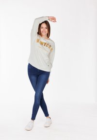 Sudadera gris claro con "NEW YORK" en letras doradas, combinada con jeans ajustados azul oscuro y zapatillas blancas, sobre un fondo blanco.