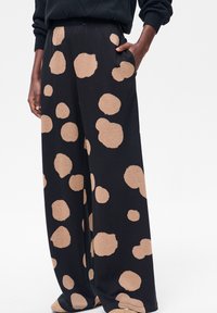 Zwarte broek met wijde pijpen en grote onregelmatige beige polka dots, gedragen met een zwarte trui, hand rustend in de zak.