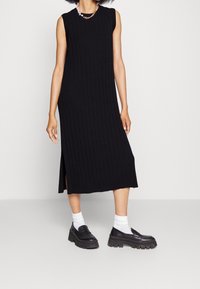 Longue robe en tricot côtelé noir, sans manches, avec une fente latérale, portée avec des mocassins noirs et des chaussettes blanches. Style et texture minimalistes.