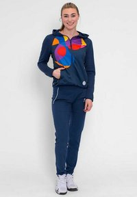 Felpa con zip blu navy con motivi geometrici colorati sul davanti, abbinata a pantaloni blu navy e scarpe da ginnastica bianche.