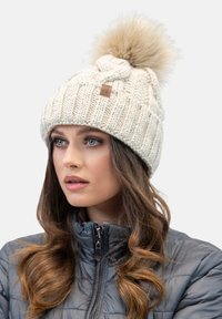 Gorro de punto crema con un pompón esponjoso de color marrón claro en la parte superior, que presenta una textura acanalada y un acento de etiqueta de cuero.