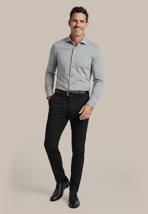 Man smiling wearing a light gray button-up shirt tucked into black dress pants with a black belt and black dress shoes.