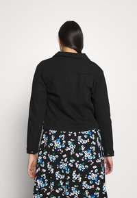 Czarna dżinsowa kurtka o dopasowanym kroju, z kołnierzykiem w kształcie punktu i mankietami na guziki. W tle widoczna spódnica w kwiatowy wzór z niebieskimi i białymi kwiatami.