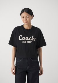 Zwarte cropped t-shirt met "Coach New York" in het wit gedrukt. Heeft een ronde halslijn en korte mouwen, gecombineerd met donkere spijkerbroek.