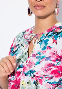 Florale Bluse mit lebhaften rosa, grünen und blauen Mustern, mit einem V-Ausschnitt und einer Bindegarnitur an der Vorderseite. Auffällige große geometrische Ohrringe.