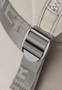 Détail de sangle grise et de boucle avec un tissu texturé, un fermoir métallique lisse et des accents cousus sur un fond beige.