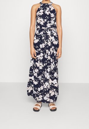 Kvinde iført en ærmeløs navyblå maxikjole med hvidt blomsterprint og bindebånd i taljen, kombineret med hvide slidesandaler.