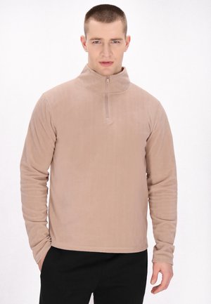 Jonge man met kort haar draagt een beige trui met een kwart ritssluiting en zwarte broek, staat met één hand in zijn zak tegen een witte achtergrond.