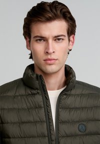 Chaqueta acolchada verde oliva con un cuello alto, con costuras horizontales, un cierre frontal de cremallera y un logotipo redondo azul marino cerca del pecho izquierdo.
