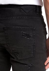 Mustat farkut denim, joissa on kulunut muotoilu useilla repeämillä ja paikoilla. Takatasku näkyvissä ja vyötärön yläpuolella brändilogo.