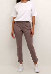 Jasnobrązowe bawełniane spodnie chino o wąskim kroju, z bocznymi kieszeniami i subtelnym detalem na krawędzi. Połączone z luźną białą koszulką i białymi tenisówkami.