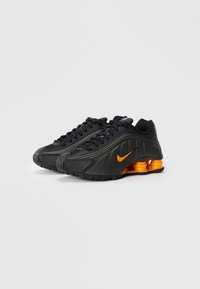 Par črnih športnih čevljev Nike z oranžnimi blažilnimi podplati in oranžnimi logotipi Swoosh, prikazanih na enobarvni svetli podlagi.