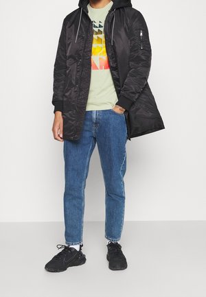 Chaqueta bomber con capucha y cremallera en negro, llevada sobre una camiseta gráfica verde claro y jeans azules, combinada con zapatillas negras.