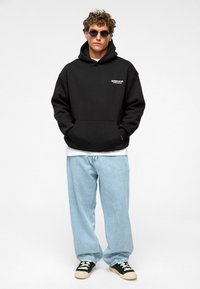 Zwarte hoodie met een voorzak en witte tekst op de borst, gecombineerd met lichtblauwe wijde jeans en zwarte sneakers met witte accenten.