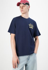 Young person wearing a red cap, navy blue graphic t-shirt with a yellow turtle and text, and light blue jeans, standing against a white background.