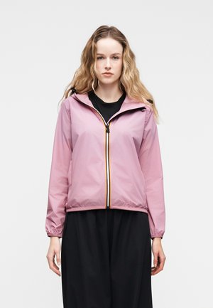 Frau trägt eine hellrosa Jacke mit Reißverschluss und elastischen Bündchen über einem schwarzen Oberteil und weiten schwarzen Hosen, steht vor einem einfarbigen hellen Hintergrund.