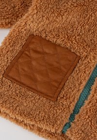 Tasca in tessuto trapuntato marrone su un capo in pile morbido color beige, con una cerniera verde visibile che corre lungo il bordo.