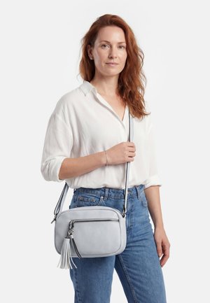 Punapäine naine, kes kannab valget särk ja siniseid teksapükse, hoides helehallis risti keha kotis, millel on tutt ja esilukk.