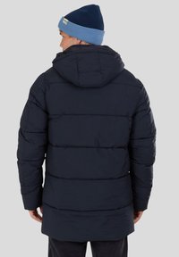 Giubbotto imbottito blu scuro con cappuccio, caratterizzato da cuciture orizzontali e un retro più lungo. Abbinato a un berretto di maglia blu chiaro.