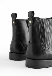 Stivali alla caviglia in pelle nera con texture rigata, accenti dorati e linguette per l'estrazione. Presentano una suola in gomma piatta e dettagli di cucitura rinforzati.