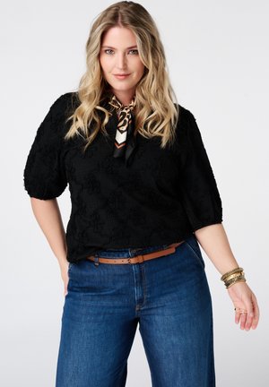 Femme blonde portant un haut noir texturé, un foulard imprimé animalier autour du cou, un jean bleu avec une ceinture marron et des bracelets dorés sur fond blanc.
