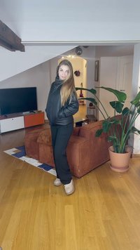 Czarna pikowana kurtka z zamkiem błyskawicznym, z wysokim kołnierzem i bocznymi kieszeniami. Czarne spodnie i beżowe buty sportowe. Przytulne wnętrze z rośliną.