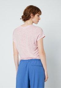 Rozā īsām piedurknēm T-krekls ar apaļu kakla izgriezumu, vilkts kopā ar zilām elastīgām biksēm. Audumā redzama viegla tekstūra. Parādīts skats no aizmugures.