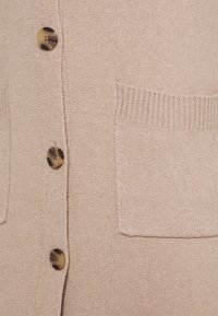 Béžový pletený kardigan s knoflíkovým zapínáním, dvěma předními kapsami a žebrovanými detaily. Měkká textura s hladkým povrchem.