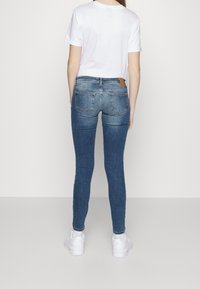 Jeans ajustados de mezclilla azul con cintura alta, que cuentan con dos bolsillos traseros y una etiqueta de cuero marrón en la cintura, combinados con una camiseta blanca.