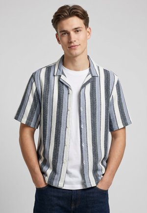 Junger Mann trägt ein kurzärmeliges Hemd mit vertikalen weißen, blauen und grauen Streifen über einem weißen T-Shirt, die Hände in den Taschen.