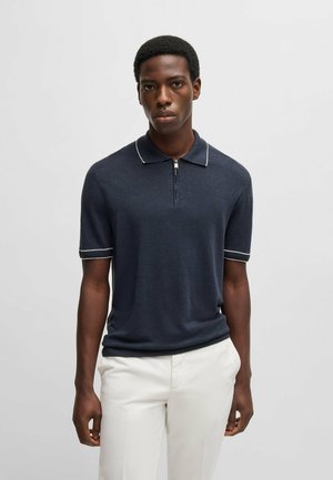 Marineblauwe polo met korte mouwen, col, ritssluiting aan de voorkant en witte piping op de kraag en mouwen; gemaakt van zachte, gestructureerde stof.