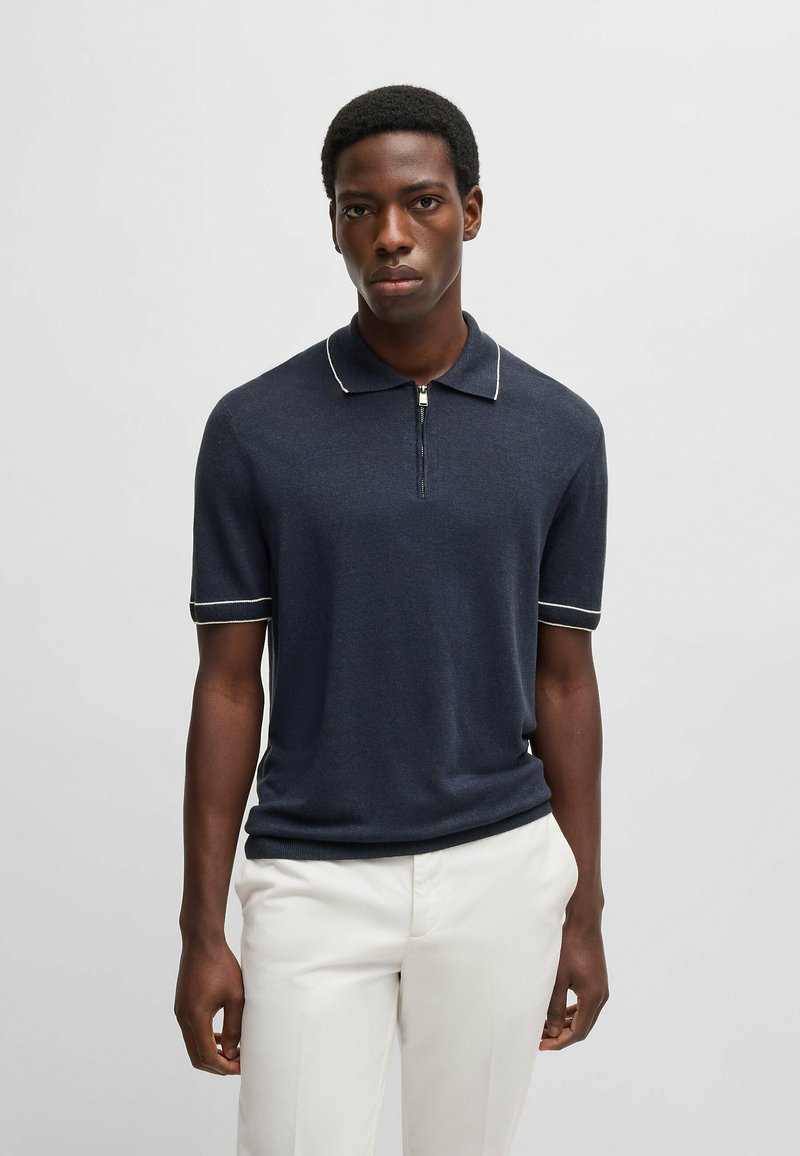 Granatowa koszulka polo z krótkim rękawem, z kołnierzykiem, zamkiem błyskawicznym z przodu oraz białymi lamówkami na kołnierzyku i rękawach; wykonana z miękkiego, teksturowanego materiału.