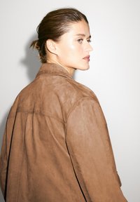 Brun ruskindjakke med løs pasform, der har en spids krave, syningsdetaljer og en glat tekstur. Modellen ses fra siden.