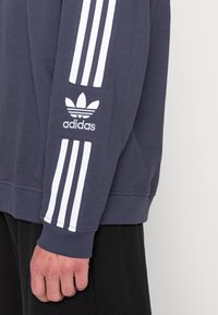 Laivaston sininen huppari, jossa on valkoinen Adidas-logo hihassa, ja hihassa on kolme valkoista raitaa sekä ribbireunat.