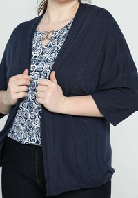 Mörkblå cardigan med ribbat mönster och vida ärmar, med en nyckelhålsglipa och metallisk detaljer. Bärs över en mönstrad topp.