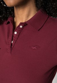 Burgundské polo tričko s límečkem, vybavené třemi bílými knoflíky a vyšitým logem na levé hrudi. Bavlněná textura.