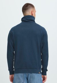 Uomo che indossa un maglione a collo alto blu scuro e jeans azzurri chiari, in piedi di spalle contro uno sfondo semplice.
