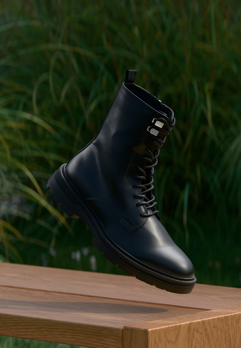 Crna kožna gležnjača s okruglim vrhom, vezicama, metalnim okicama i teksturiranim potplatom, ovješena na zelenoj pozadini.