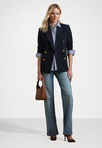 Dubbelzijdig marineblauw blazer met gouden knopen, gecombineerd met een gestreept overhemd, lichtblauwe flared jeans en bruine hoge hakken, terwijl ze een bruine handtas vasthoudt.