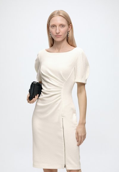 Femme aux longs cheveux blonds portant une robe blanche froncée et tenant une petite pochette noire sur un fond blanc uni.