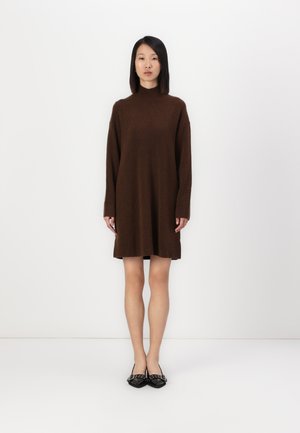 Braunes Strickkleid mit hohem Kragen, langen Ärmeln und lässiger Passform, kombiniert mit schwarzen flachen Schuhen mit dekorativer Hardware.