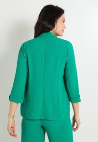 Chaqueta verde texturizada con cuello, mangas tres cuartos y puños arremangados. Presenta un corte recto y detalle de costura en la parte posterior.